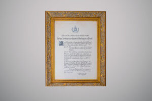 Visão frontal e completa da cópia do decreto imperial que cria os cursos de odontologia no Brasil, datado de vinte e cinco de outubro de mil oitocentos e oitenta e quatro. O documento está centralizado em uma moldura dourada com entalhes decorativos. No topo, há o brasão do Império do Brasil.