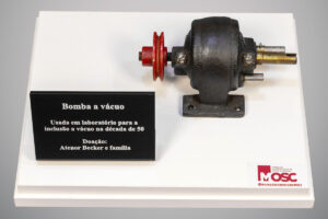 Fotografia superior frontal de uma bomba a vácuo de ferro fundido escuro com polia vermelha, montada sobre base branca. À esquerda, uma placa informa que o equipamento foi usado em laboratório na década de cinquenta para inclusão a vácuo. A peça foi doada por Atenor Becker e família ao Museu da Odontologia de Santa Catarina.