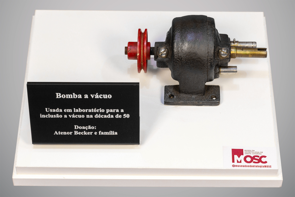Fotografia superior frontal de uma bomba a vácuo de ferro fundido escuro com polia vermelha, montada sobre base branca. À esquerda, uma placa informa que o equipamento foi usado em laboratório na década de cinquenta para inclusão a vácuo. A peça foi doada por Atenor Becker e família ao Museu da Odontologia de Santa Catarina.