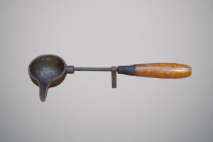 Utensílio antigo de ferro com cabo de madeira, formado por uma pequena concha arredondada com bico para despejar líquidos.