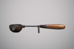 Utensílio antigo de ferro com cabo de madeira, formado por uma pequena concha arredondada com bico para despejar líquidos.