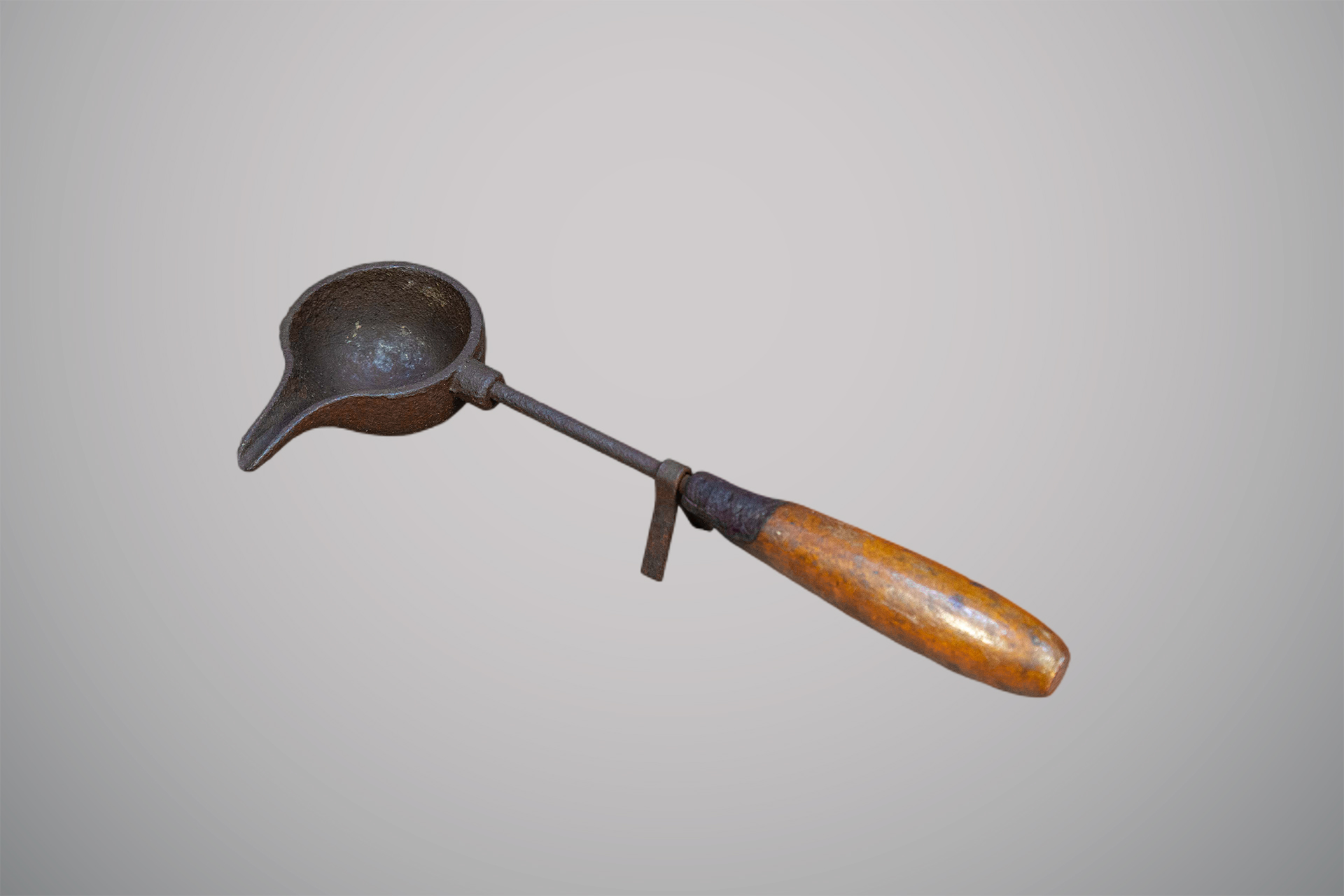 Utensílio antigo de ferro com cabo de madeira, formado por uma pequena concha arredondada com bico para despejar líquidos.