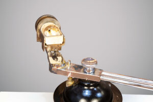 Close em parte superior de uma centrífuga Stif da década de cinquenta, mostrando detalhes metálicos em tom prateado e dourado, com suporte usado na fundição de ligas odontológicas.