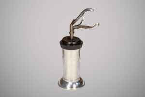 Instrumento médico antigo de metal prateado com base cilíndrica e alavanca superior em formato de pinça.