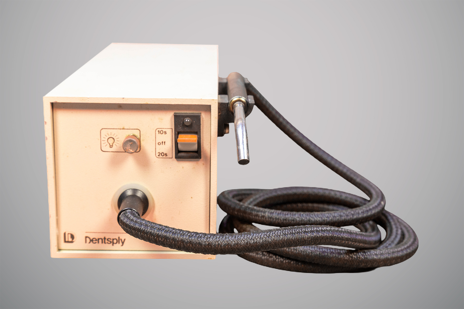 Equipamento odontológico retangular da marca Dentsply, de cor bege, com cabo grosso preto ligado a uma peça metálica cilíndrica. Na frente há um botão prateado com símbolo de lâmpada, um seletor com as opções “dez segundos”, “off” e “vinte segundos”, além de uma chave laranja de liga e desliga.