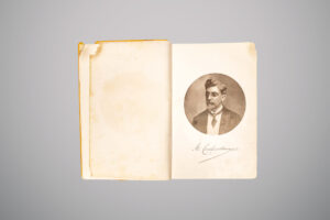 Livro antigo aberto, com página esquerda em branco e sinais de desgaste, e página direita exibindo retrato oval em preto e branco de um homem de óculos, barba e terno, acompanhado de assinatura manuscrita abaixo.