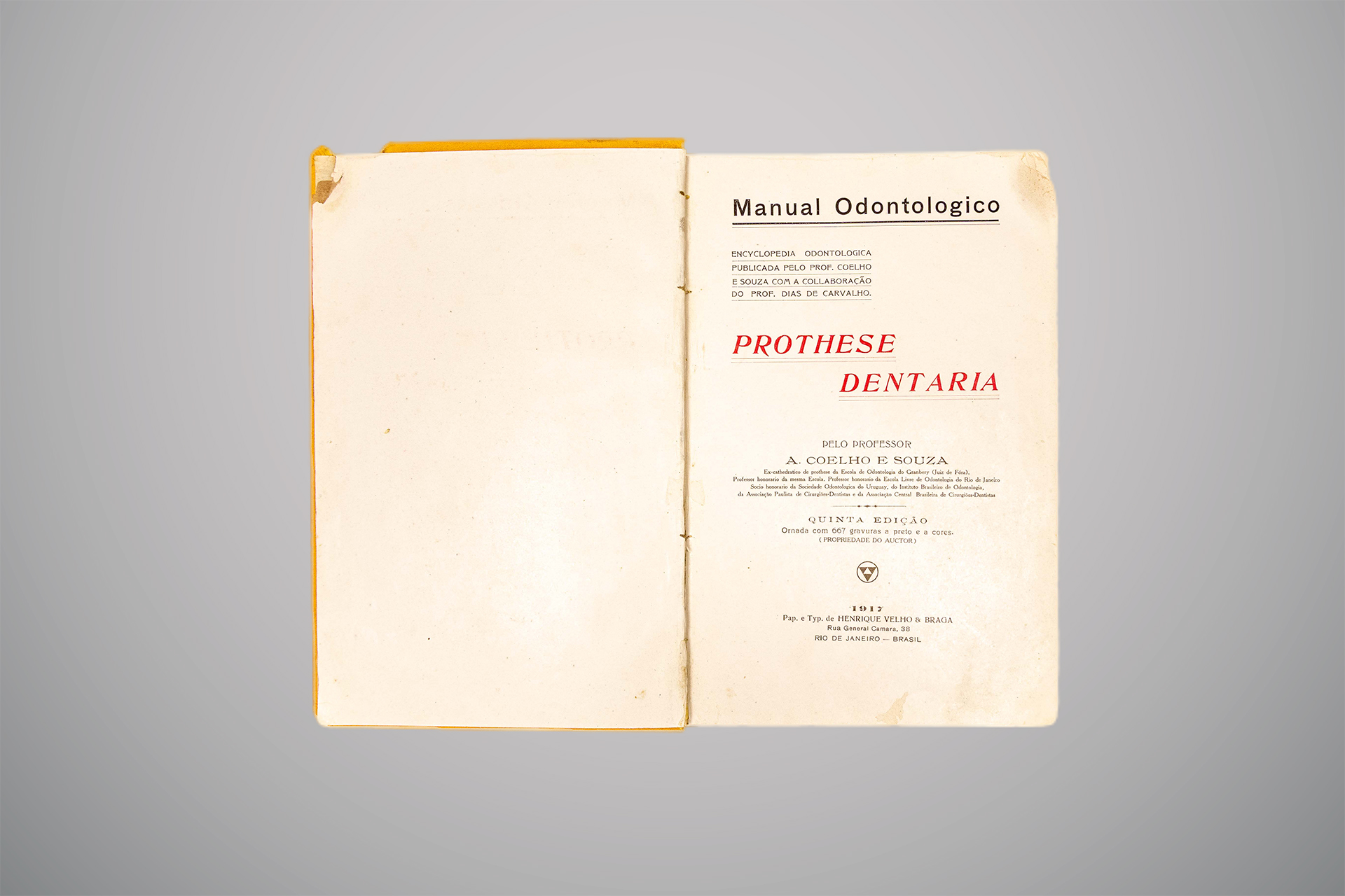 Livro antigo aberto, com página esquerda em branco e página direita apresentando a capa interna do título “Manual Odontologico – Prothese Dentaria”, escrito em letras pretas e vermelhas, com informações do autor A. Coelho e Souza, detalhes editoriais e data de mil novecentos e dezessete.