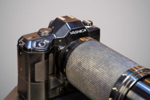 Câmera fotográfica preta da marca Yashica em close, com lente longa e revestida por textura metálica.