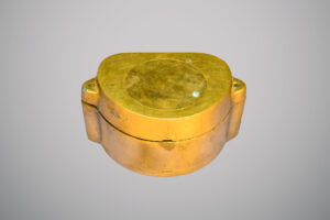Objeto metálico de cor dourada em formato arredondado, com tampa superior circular fixada por parafusos.