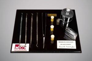 Conjunto de instrumentos odontológicos em exibição sobre base preta, com quatro espátulas metálicas, uma pinça, três frascos pequenos com tampas contendo pequenas esferas douradas, recipiente metálico com corrente e isqueiro cromado.
