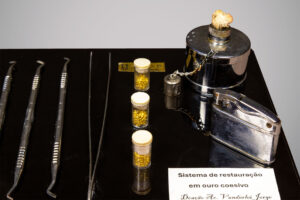 Conjunto de instrumentos odontológicos em exibição sobre base preta, com quatro espátulas metálicas, uma pinça, três frascos pequenos com tampas contendo pequenas esferas douradas, recipiente metálico com corrente e isqueiro cromado.