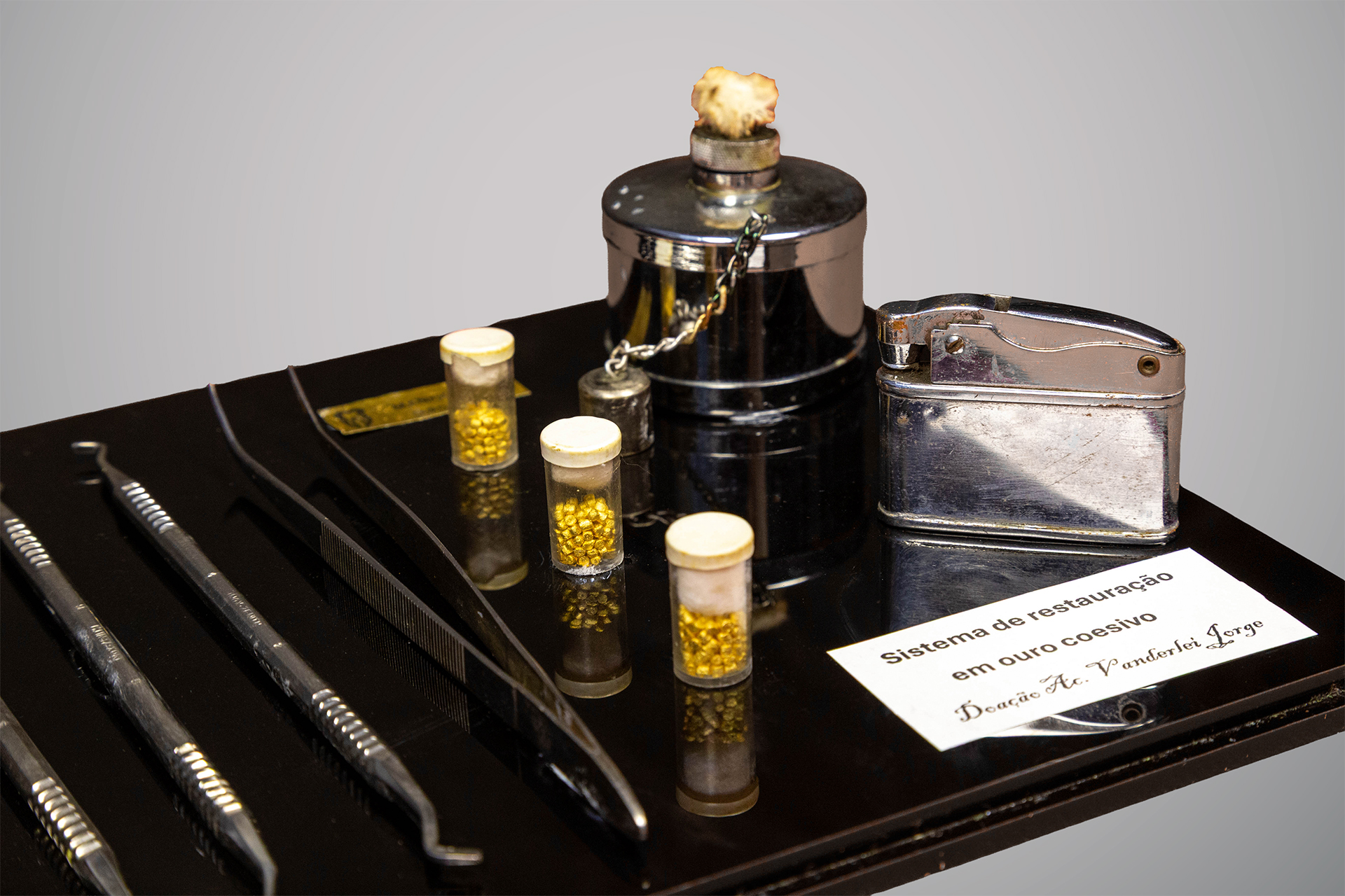 Conjunto de instrumentos odontológicos em exibição sobre base preta, com quatro espátulas metálicas, uma pinça, três frascos pequenos com tampas contendo pequenas esferas douradas, recipiente metálico com corrente e isqueiro cromado.